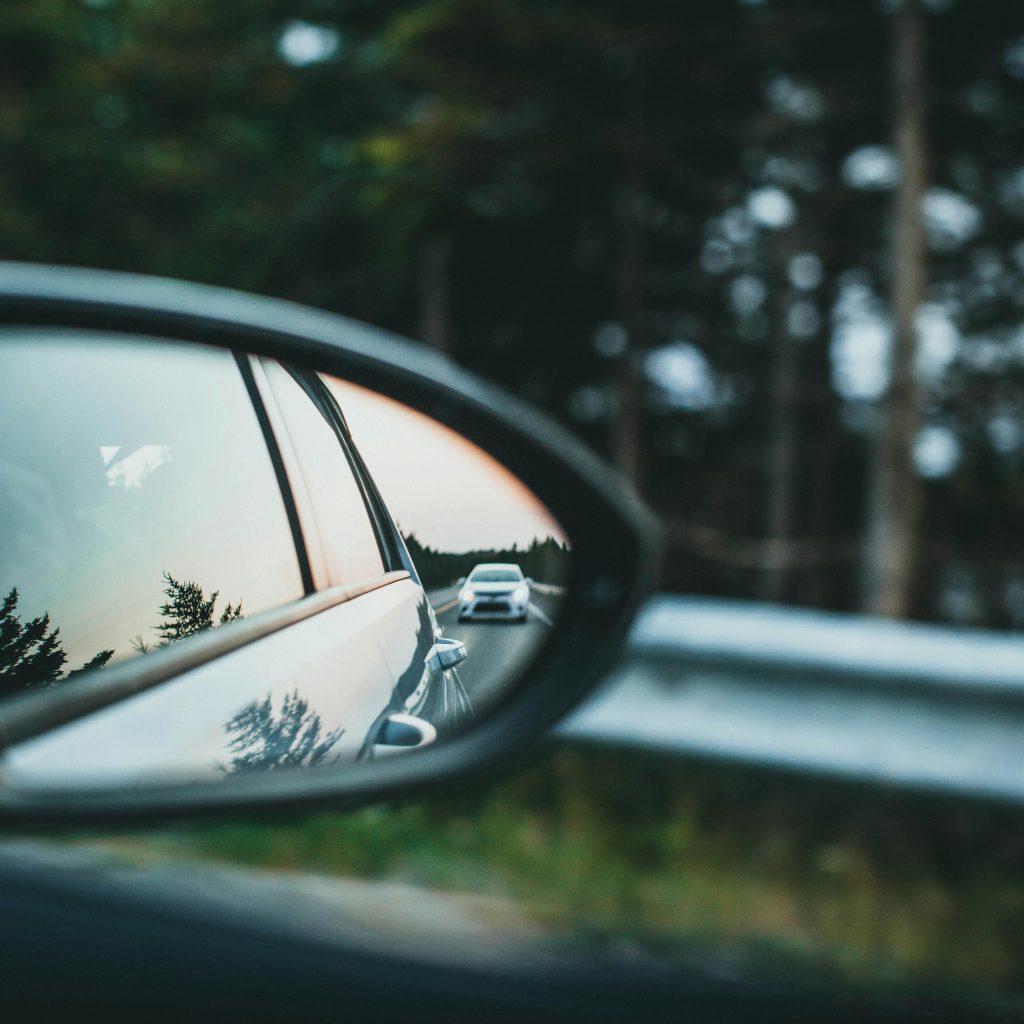 A car is reflected in a side mirror on a scenic forest road.